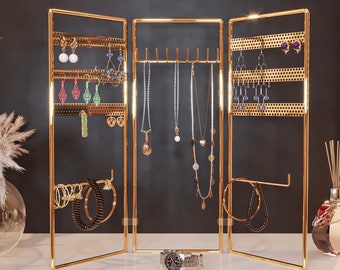 May include: A gold-colored, three-panel jewelry organizer with multiple earring and necklace displays. The organizer is filled with various earrings, necklaces, and bracelets. A silver wristwatch sits in front of the organizer.