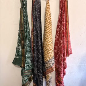 May include: Four rectangular scarves hanging from a horizontal string. The scarves feature various patterns in green, black, yellow, and red. Each piece displays intricate block-printed designs and finished edges with thin, contrasting colored borders.
