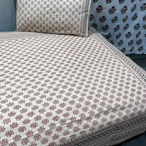 May include: A bed sheet and matching pillowcase set featuring a repeating small floral pattern in dark red on a cream background. The edges are finished with a black and white geometric zigzag border, providing a structured frame to the design.