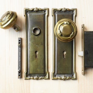 May include: A set of antique brass door hardware, including a pair of door plates, two door knobs, a strike plate, and a latch. The hardware has a traditional design with ornate details.