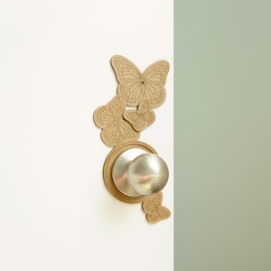 Könnte beinhalten: Ein silberner Türknauf mit einem dekorativen Holzschmetterlingsdesign drumherum. Die Schmetterlinge sind in Brauntönen gehalten und an einer weißen Tür befestigt.