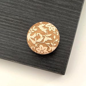 May include: A circular wooden button featuring a floral pattern. The button is light brown and intricately carved with flowers and foliage. It rests on a dark grey wooden surface.