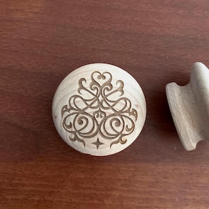 Peut inclure: Un bouton de meuble en bois clair avec un motif décoratif gravé de volutes, de cœurs et d'une étoile. Le bouton est rond et a une finition en bois naturel. Un autre objet en bois est partiellement visible.