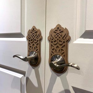 Könnte beinhalten: Zwei Türgriffe mit dekorativen Holzauflagen. Die Auflagen sind hellbraun und weisen ein kompliziertes, durchbrochenes Design auf. Die Türgriffe sind silberfarben und gebogen. Die Türen sind weiß.