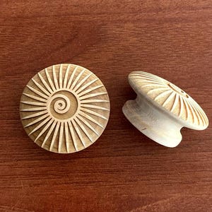 May include: Two wooden cabinet knobs with a sunburst design. One knob is lying flat, and the other is on its side. The knobs are light-coloured wood with a spiral centre and radiating lines.