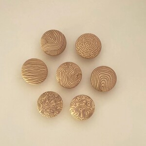 Op de afbeelding: Zeven ronde houten knopen gerangschikt in een groep. Elke knoop heeft een uniek, met laser gegraveerd patroon, waaronder topografische lijnen, concentrische cirkels en gestippelde texturen. Het hout heeft een lichte, natuurlijke beige afwerking.