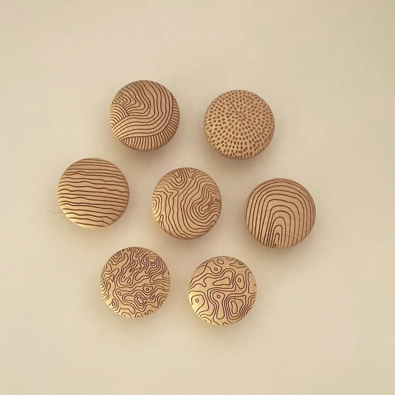 Op de afbeelding: Zeven ronde houten knopen gerangschikt in een groep. Elke knoop heeft een uniek, met laser gegraveerd patroon, waaronder topografische lijnen, concentrische cirkels en gestippelde texturen. Het hout heeft een lichte, natuurlijke beige afwerking.