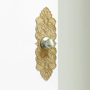 May include: A decorative door knob plate with a detailed, intricate design in a light brown colour. The plate is attached to a white door and features a silver door knob.