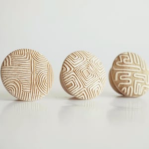 Op de afbeelding: Drie ronde, beige keramische kastknoppen met witte geometrische patronen. De knoppen zijn in een rij gerangschikt, elk met een uniek ontwerp van lijnen en vormen. De knoppen zijn waarschijnlijk voor meubels.