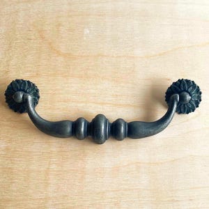 May include: Antique bronze drawer pull with a floral design on each end. The pull has a curved handle with decorative details. The hardware is set against a light wood surface.