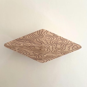 May include: A diamond-shaped wooden wall hook featuring a light tan natural wood grain finish and a dark brown laser-engraved topographic map pattern consisting of irregular, flowing contour lines across the entire front surface of the object.