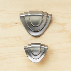 May include: Two silver-toned cabinet knobs with an art deco design. The knobs are shaped like a rounded triangle with three concentric arches.