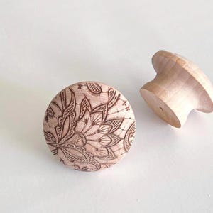May include: Two unfinished wooden cabinet knobs. One is round with a detailed floral design etched into the surface. The other knob is a mushroom shape. Both are light brown in color and sit on a white background.