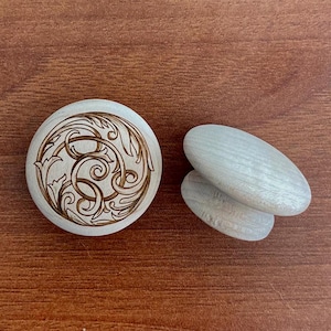 May include: A wooden round object with an engraved design and two oval-shaped wooden objects. The round object has a detailed, swirling pattern. The oval objects are smooth and light-colored. All objects are on a brown surface.
