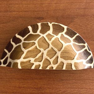 May include: A wooden, semi-circular cabinet pull with a giraffe pattern. The pull features a light brown background with darker brown and cream-coloured irregular shapes, resembling a giraffe's coat. The pull is set against a wooden surface.