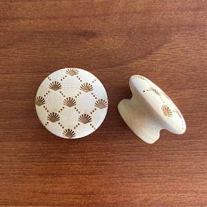 May include: Two wooden cabinet knobs. One is round, the other is a pull. Both are off-white with a repeating pattern of small, stylized sunbursts in a grid, connected by dotted lines. The knobs are on a brown wooden surface.