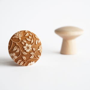Puede incluir: Perilla de armario redonda de madera marrón con un estampado floral. La perilla presenta un diseño detallado de hojas y flores. Una segunda perilla de madera sin terminar y borrosa está en el fondo. Las perillas son probablemente para muebles.