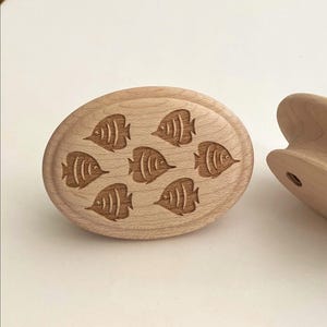 Peut inclure: Un objet en bois de couleur brun clair de forme ovale présentant un motif gravé au laser de sept petits poissons disposés en groupe. La surface montre une texture de grain de bois naturel, avec un bord arrondi lisse et un profil latéral visible.