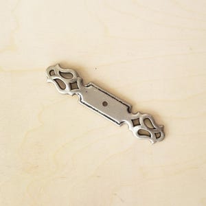 May include: Silver metal drawer pull with an ornate design. The pull has a rectangular centre with a hole for a screw.