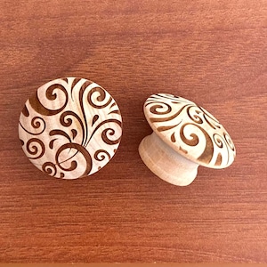 May include: Two wooden cabinet knobs with a decorative swirl design. One knob is flat, and the other is mushroom-shaped. The knobs are light-colored wood with a dark brown swirl pattern. They are set against a brown wooden surface.