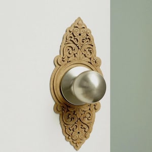 Peut inclure: Un bouton de porte décoratif avec une finition en métal brossé. Le bouton est encastré dans une plaque arrière en bois marron clair, ornée et détaillée. La plaque arrière présente un motif symétrique de volutes. Le bouton est monté sur un mur blanc.