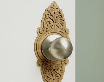 May include: A door knob with a smooth, brushed silver finish centered on a decorative, light brown wooden backplate. The backplate features an intricate, symmetrical carved scrollwork pattern that extends above and below the central knob.