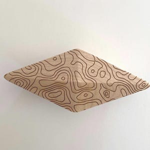 May include: A diamond-shaped wooden object with a light tan natural wood grain finish. The surface is decorated with dark brown, thin, continuous lines that form an intricate, irregular topographic map pattern across the entire front face.