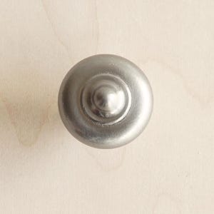 May include: A close-up of a brushed nickel cabinet knob. The round knob has a slightly raised centre and a matte finish. The hardware is designed for furniture and home decor.