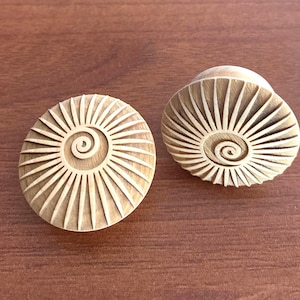 Könnte beinhalten: Zwei hellbraune Holz-Ohrstecker mit einem geschnitzten Spiral- und Strahlenmuster, das an eine Nautilusmuschel erinnert. Die runden Ohrstecker werden auf einer Holzoberfläche präsentiert. Dekorativer Artikel.