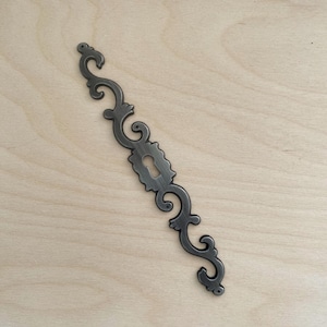 Puede incluir: Una placa de metal oscuro para cerradura con un diseño alargado y simétrico de volutas y una abertura central para la llave. El herraje tiene un acabado cepillado e incluye pequeños orificios circulares para el montaje.