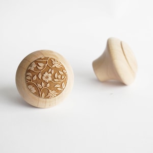 Könnte beinhalten: Zwei hölzerne Schrankknöpfe. Einer ist rund mit einem floralen Design, das in die Oberfläche geschnitzt ist. Der andere hat eine glatte, abgerundete Form. Beide sind aus hellem Holz und vor weißem Hintergrund.