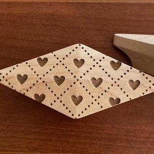 Könnte beinhalten: Dekoratives Holzelement in Diamantform mit Herz-Ausschnitten und gepunkteten Linien. Das hellfarbene Holz kontrastiert mit dem dunkelbraunen Hintergrund. Ein zweites, unfertiges Holzstück ist sichtbar.