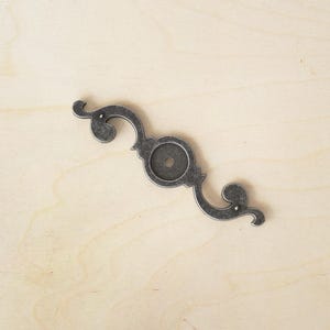 Può includere: Un pezzo di ferramenta decorativa in metallo argentato con un centro circolare e volute ornate su entrambi i lati.