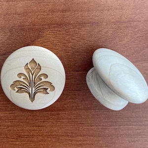 Könnte beinhalten: Zwei Holzschrankknöpfe und ein runder Holzschubladengriff. Der Schubladengriff hat ein geschnitztes Blumendesign. Die Knöpfe sind oval und glatt. Alle Artikel sind helles Holz.