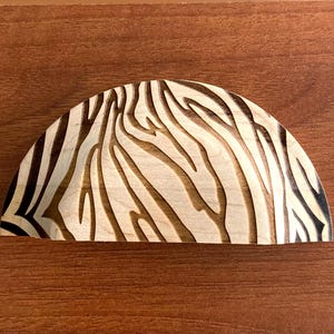 May include: A semi-circular wooden decorative item featuring a zebra stripe design. The stripes are dark brown on a light wood base. It rests on a brown wooden surface.