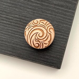 May include: A round wooden cabinet knob with an engraved swirling floral design. The knob is light brown and sits on a dark gray surface. The design is carved into the wood, creating a textured effect.