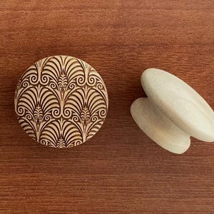 Könnte beinhalten: Ein runder Holzschrankknopf mit Art-Deco-Design und ein Paar hellfarbige Holzschubladengriffe. Der Knopf hat ein detailliertes, geätztes Muster in Braun auf hellem Holzgrund. Die Griffe sind oval geformt.