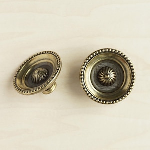 May include: Two gold-toned cabinet knobs with a decorative, raised, circular design in the center. The knobs have a beaded edge.
