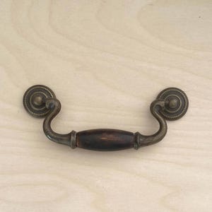 Peut inclure: Une poignée de tiroir composée d'une partie centrale en bois brun foncé, encadrée par des bras métalliques courbés au fini laiton antique. La poignée se fixe via deux plaques circulaires perlées, chacune dotée d'un petit bouton central.