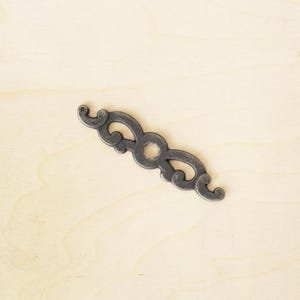 Könnte beinhalten: Dekorative, verzierte Schubladengriff aus dunkelgrauem Metall. Der Griff hat ein symmetrisches Design mit wirbelnden Details und einer zentralen kreisförmigen Öffnung. Zur Montage an Schubladen oder Schränken.