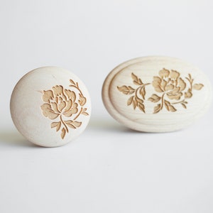 May include: Two wooden stamps with a floral design. The stamps are carved with a delicate floral pattern, featuring a single flower with leaves and stems. The stamps are made of light-colored wood and have a smooth, polished finish.