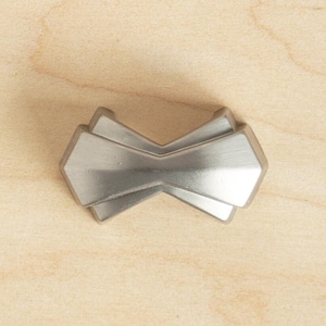 Może przedstawiać: Srebrny, geometryczny uchwyt do szafki o nowoczesnym designie. Uchwyt jest wykonany z metalu i ma gładkie, polerowane wykończenie.