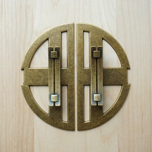 Puede incluir: Un par de tiradores de puerta de armario de latón antiguo con un diseño circular y un patrón en forma de cruz. Los tiradores están separados por un pequeño espacio.