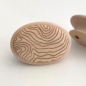 Può includere: Un pomello per mobili ovale in legno chiaro con un motivo a mappa topografica inciso al laser di linee concentriche sulla parte anteriore. Il pomello ha una finitura naturale liscia e un foro di montaggio visibile sul lato della base.