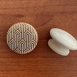Peut inclure: Un bouton de meuble rond doré avec un motif géométrique, à côté d'une paire de boutons de meuble ovales blancs. Les boutons sont posés sur une surface en bois brun chaud.