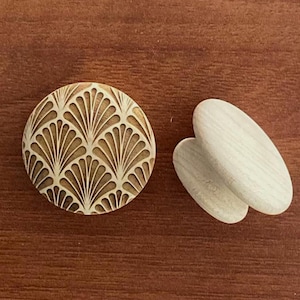 Könnte beinhalten: Ein hellbraunes, rundes Holzobjekt mit einem fächerförmigen Muster und zwei ovalen Holzobjekten. Sie liegen auf einer dunkelbraunen Holzoberfläche. Das runde Objekt hat ein detailliertes, geschnitztes Design.