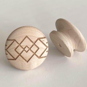 Op de afbeelding: Twee onafgewerkte houten kastknoppen. Eén is rond met een geëtst geometrisch ontwerp. De andere is een paddenstoelvormige knop. Beide zijn gemaakt van lichtgekleurd hout.