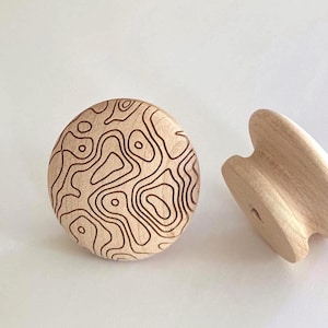 Puede incluir: Dos pomos redondos de madera con un acabado de veta natural en color tostado claro. El pomo frontal presenta un patrón de mapa topográfico grabado con láser en marrón oscuro que consiste en líneas de contorno irregulares y fluidas.