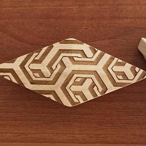 Könnte beinhalten: Ein rautenförmiges Holzeinlegearbeit mit einem geometrischen Muster aus ineinandergreifenden Formen. Die Einlage ist hellbraun mit dunkleren geätzten Linien, vor einem rötlich-braunen Holzgrund. Ein helles Holzstück befindet sich daneben.