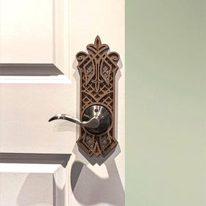 Op de afbeelding: Een decoratieve houten deurplaat met een sierlijk, geometrisch ontwerp. De plaat is gemonteerd op een witte deur en heeft een zilverkleurige deurklink. Het ontwerp heeft een warme bruine kleur en de deurklink is gebogen.